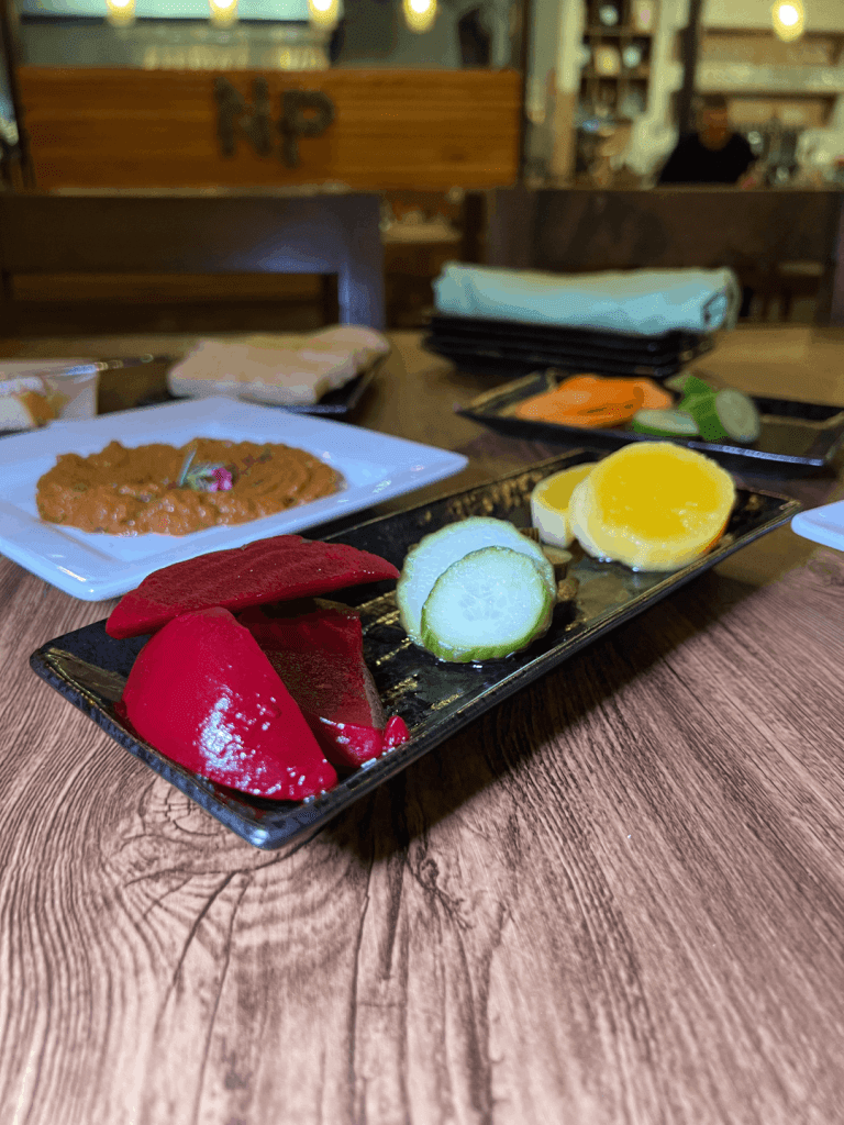 A plate with pickled vegetables, including red beets, cucumber, and yellow squash, on a wooden table with other Middle Eastern dishes in the background.