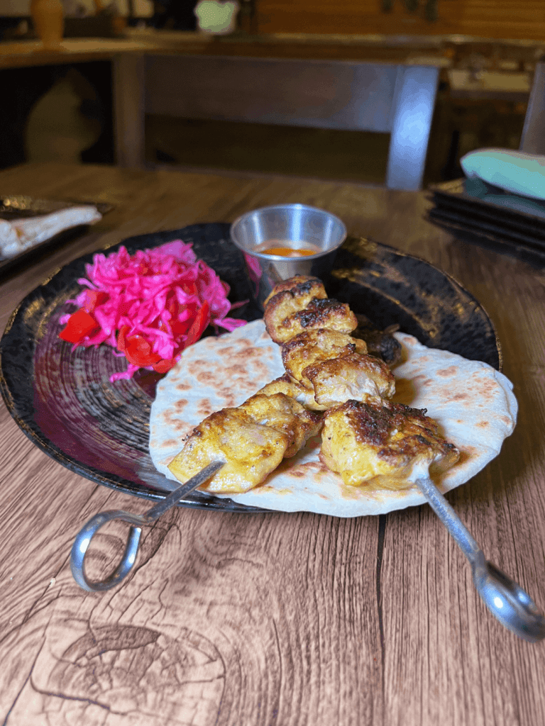 Grilled skewered chicken on a flatbread with pickled red cabbage and a small cup of sauce on a black plate.