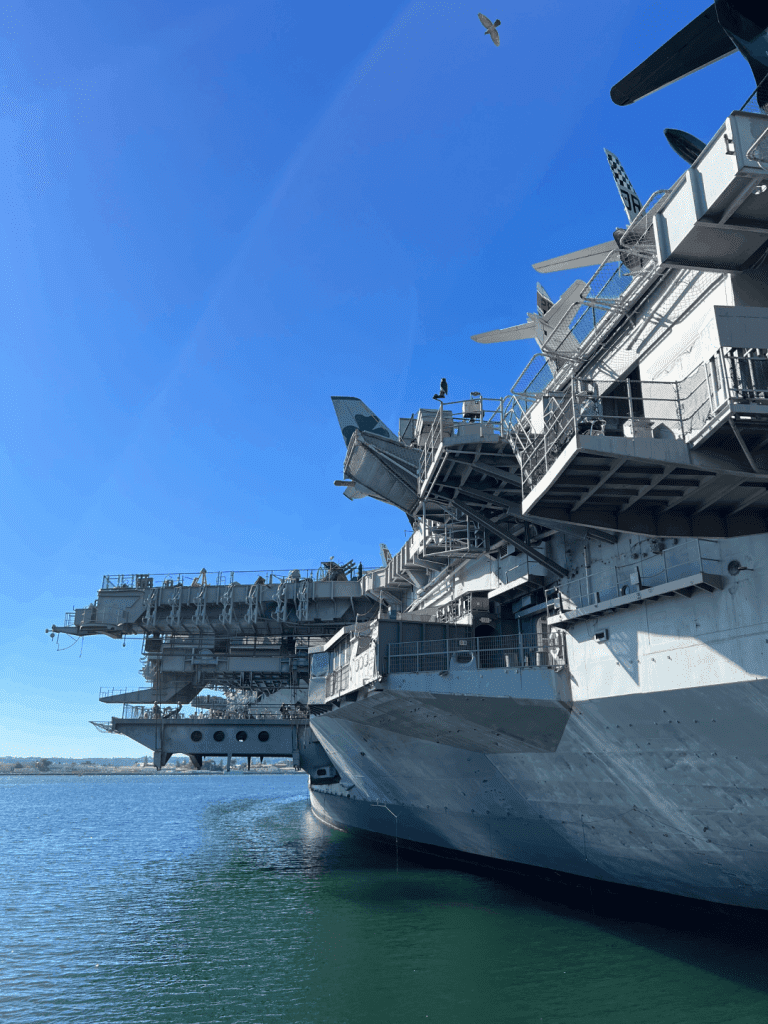 A large aircraft carrier docked in a harbor, with several aircraft on its deck and a clear blue sky overhead.