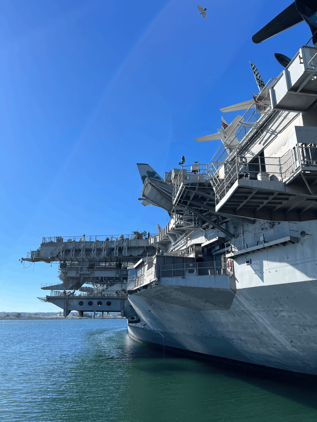 A large aircraft carrier docked in a harbor, with several aircraft on its deck and a clear blue sky overhead.