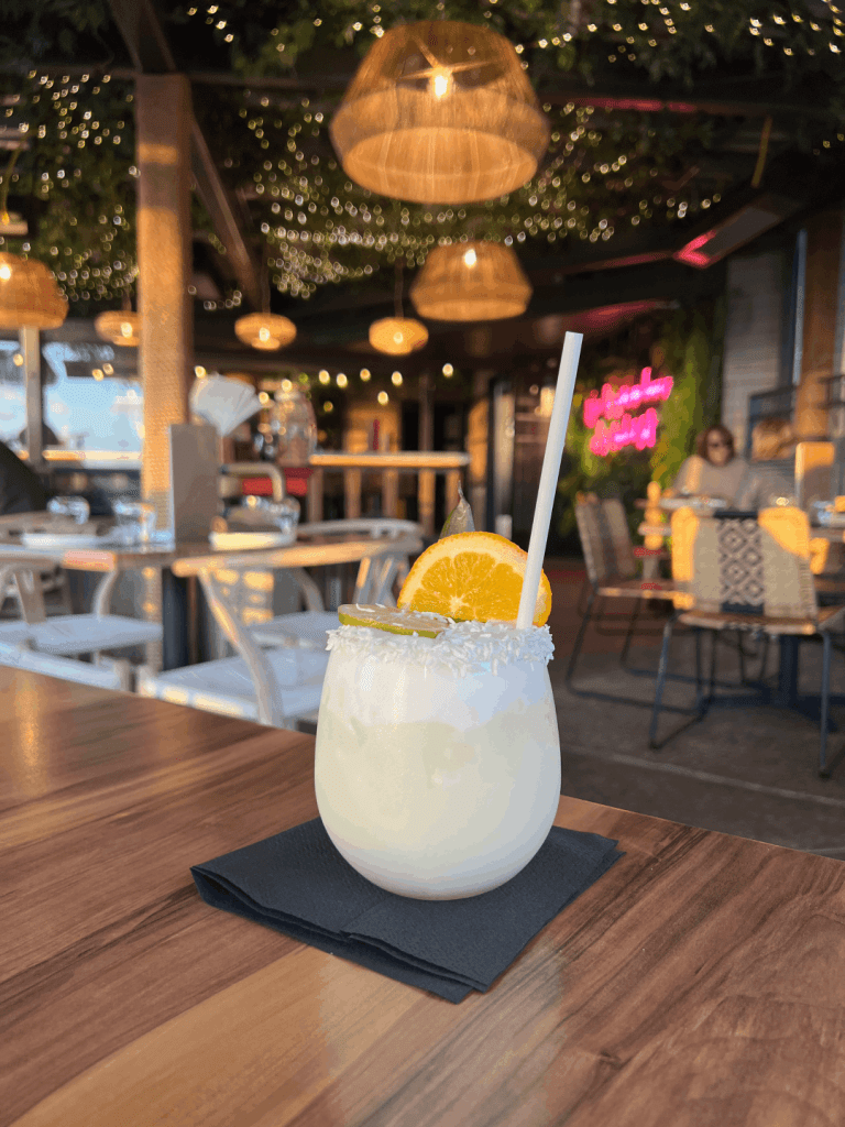 A creamy cocktail with a straw, garnished with an orange slice and lime wedge, served on a wooden table in a warmly lit restaurant with hanging lights.