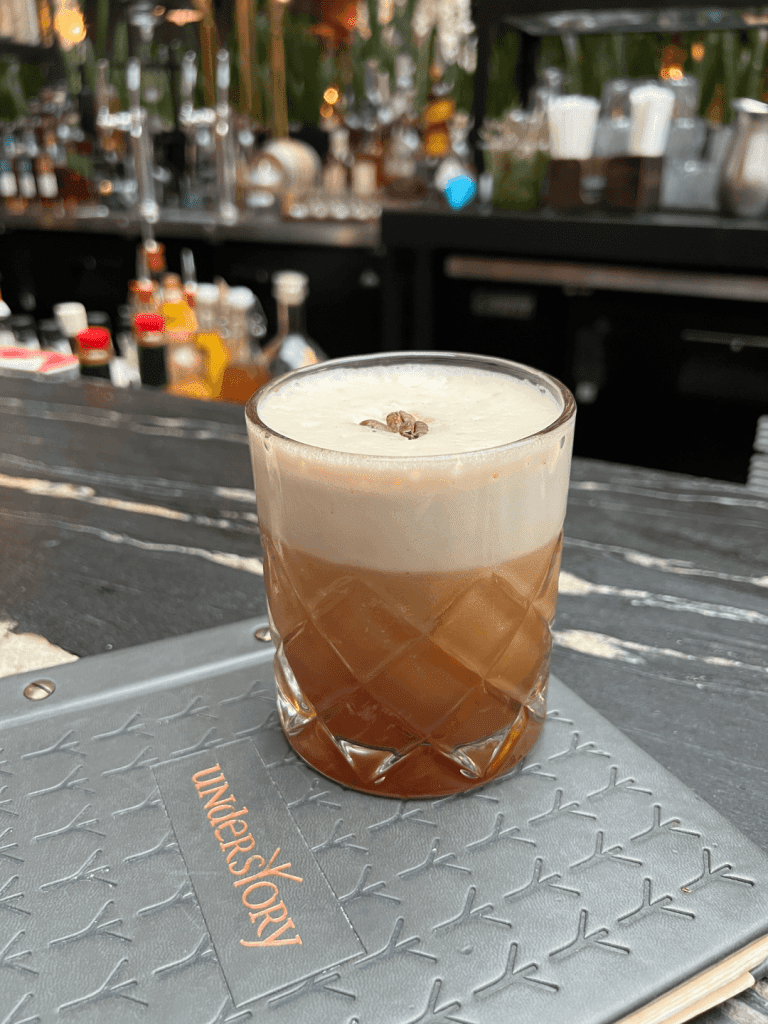 A cocktail with foam and coffee beans on top in a textured glass on a table, with a bar in the background.