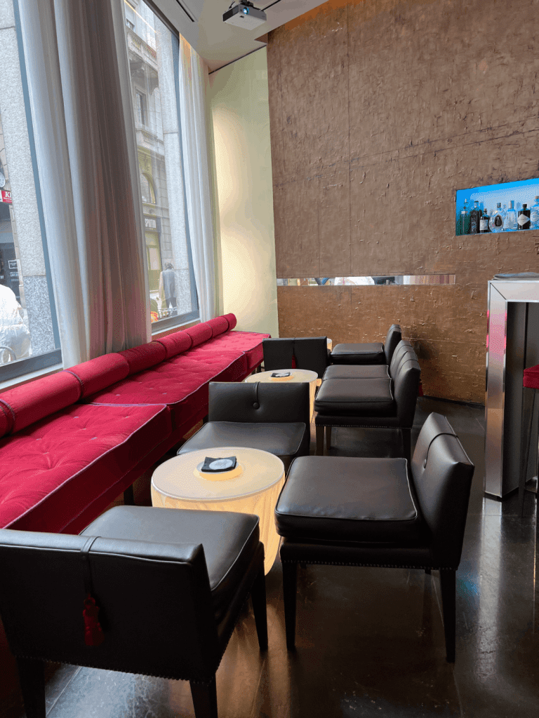 Modern lounge interior with red cushioned bench seating, black upholstered chairs, small tables, and a wall display of bottles.