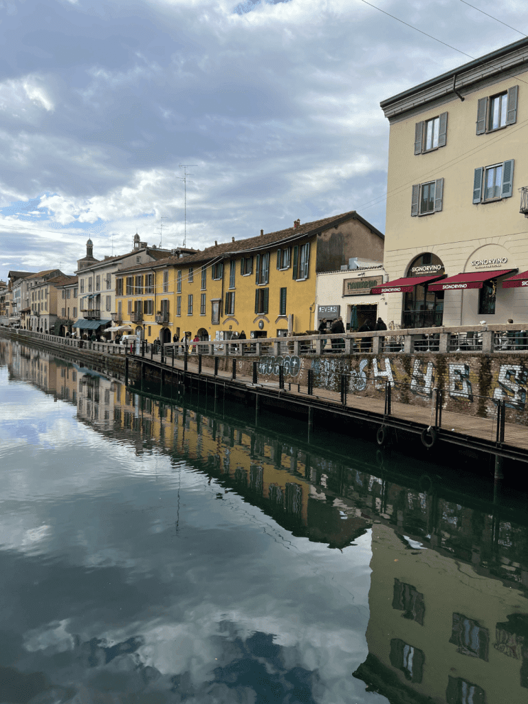 Canal-side street with colorful old buildings reflected in the water, under a cloudy sky.