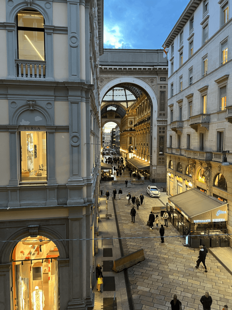 A view of Galleria Vittorio Emanuele II in Milan with people walking on the street and shops illuminated in the evening.