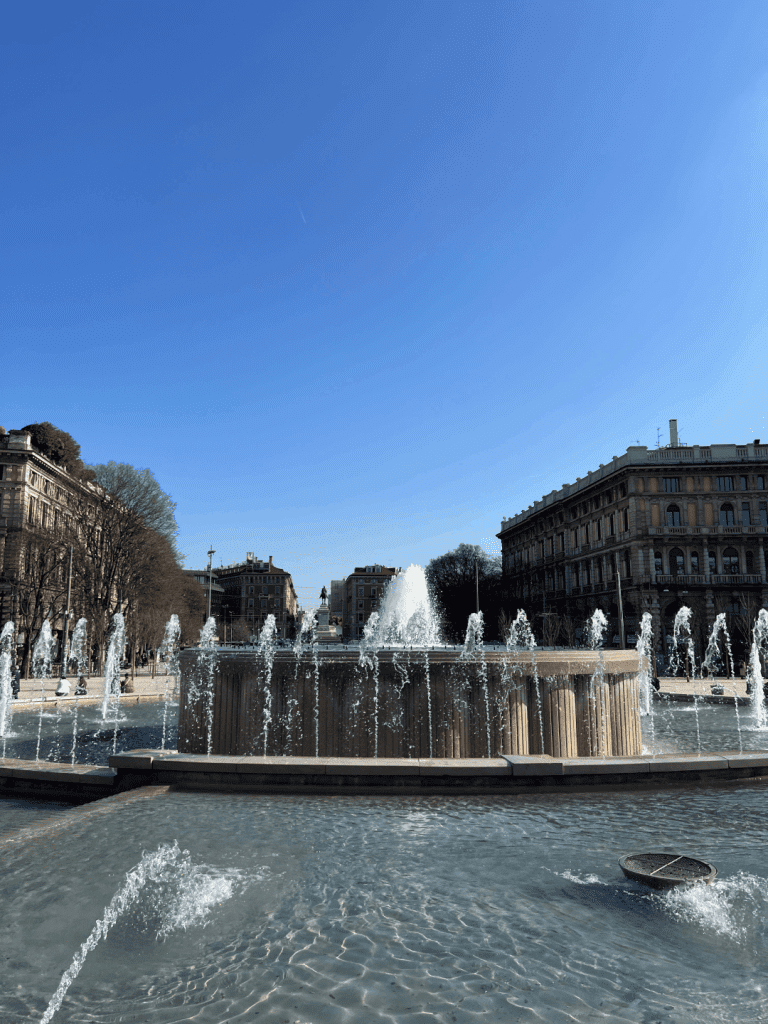 Fountain with multiple water jets in a plaza surrounded by historic buildings under a clear blue sky.