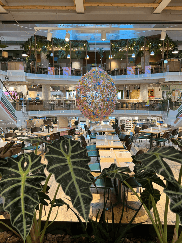 Interior of a multi-level marketplace with a hanging floral sculpture, surrounded by dining tables and large green plants.