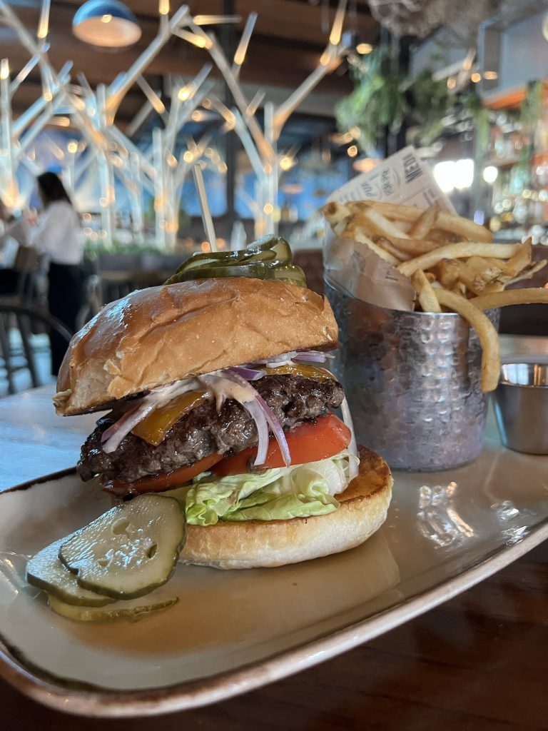 Burger with pickles, lettuce, tomato, onion, and cheese on a toasted bun, alongside a metal cup of fries on a plate, in a restaurant setting.