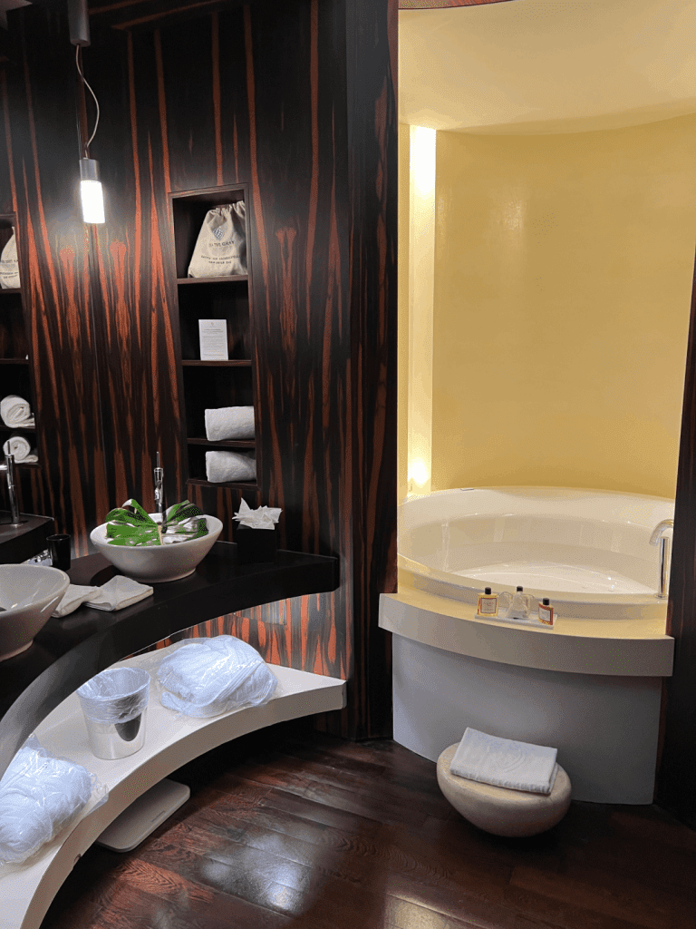 Luxurious bathroom with a large circular bathtub, dark wood paneling, and modern fixtures, featuring towels and amenities neatly arranged on shelves.