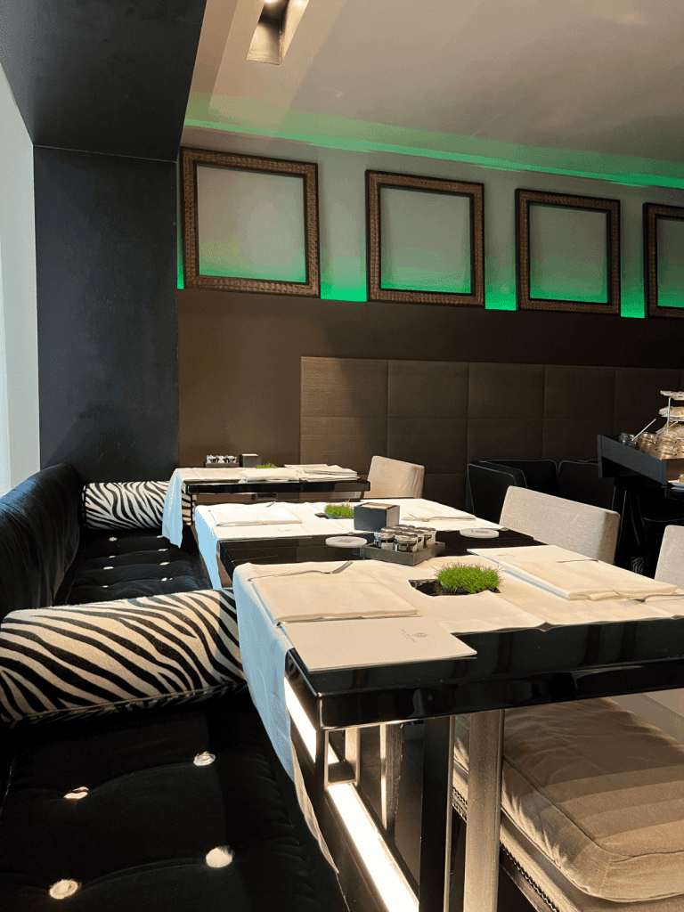 Elegant restaurant interior with a black cushioned bench, zebra-patterned pillows, a sleek black table set with white napkins, chairs, and modern decor elements under soft green lighting.