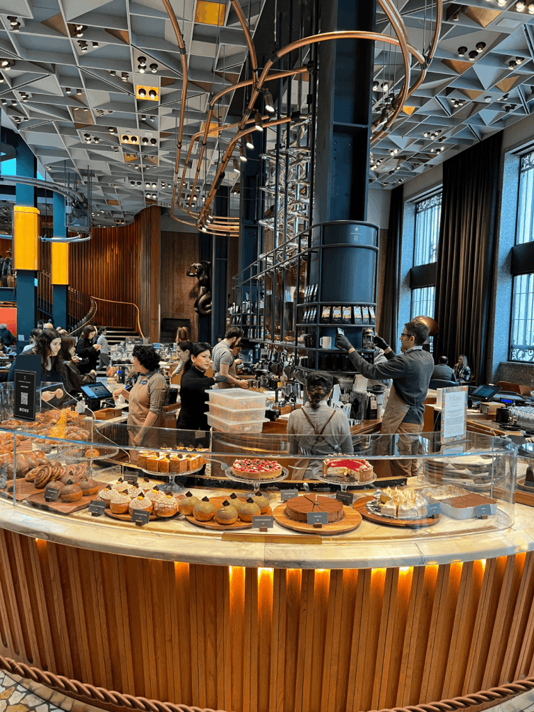 A bustling cafe with a modern industrial design, featuring a pastry display filled with a variety of cakes and baked goods. Customers and staff are engaged at the counter under a unique geometric ceiling.