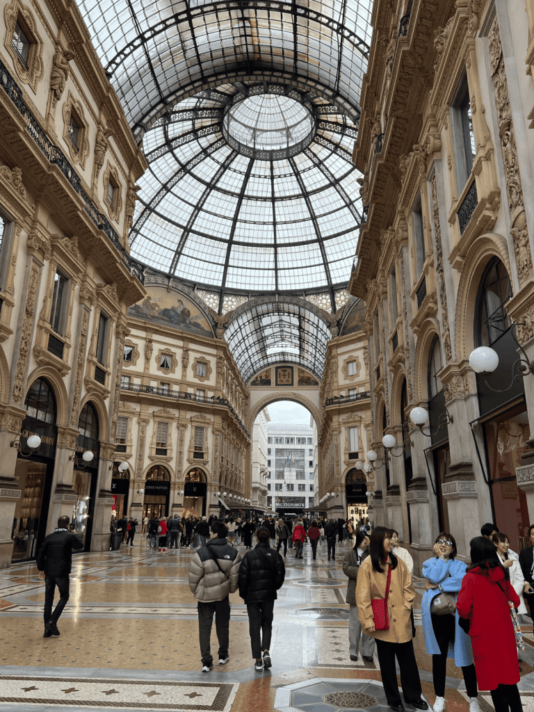 Interior of Galleria Vittorio Emanuele II in Milan, with crowds walking under its arched glass and iron roof.