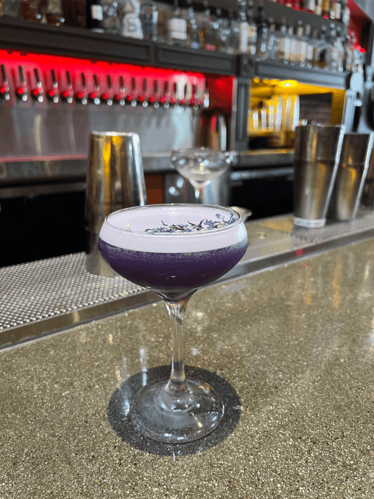 A purple cocktail in a coupe glass, garnished with dried flowers, on a bar countertop with shakers and bottles in the background.