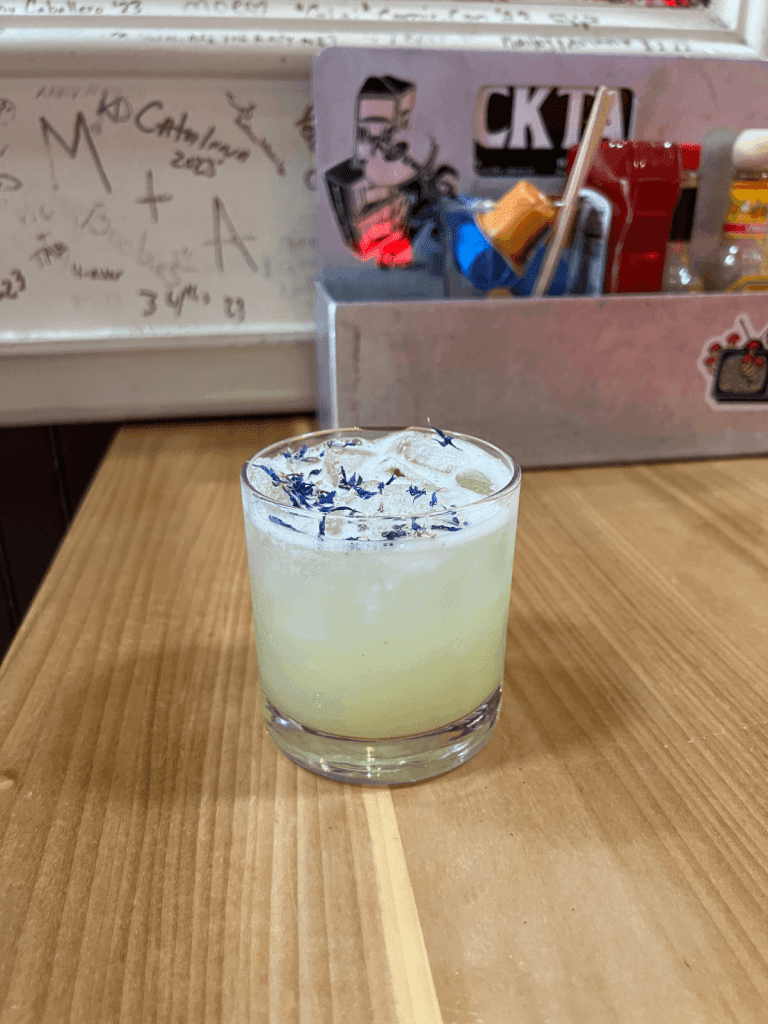 A glass of pale yellow drink with ice and purple flower petals on top, placed on a wooden table.