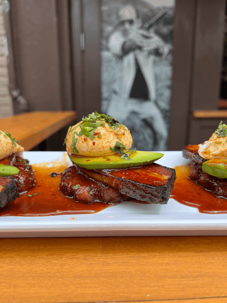 A dish featuring glazed meat topped with avocado slices and a dollop of seasoned cream, garnished with herbs, on a white plate.