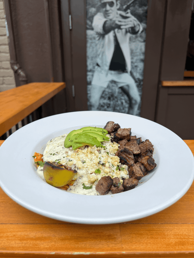 A plate of grilled steak cubes, avocado slices, roasted corn, and a grilled lime wedge on a wooden table.