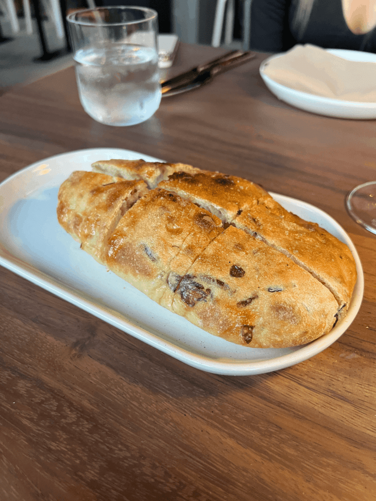 A loaf of sliced bread with olives on a plate, next to a glass of water on a wooden table.