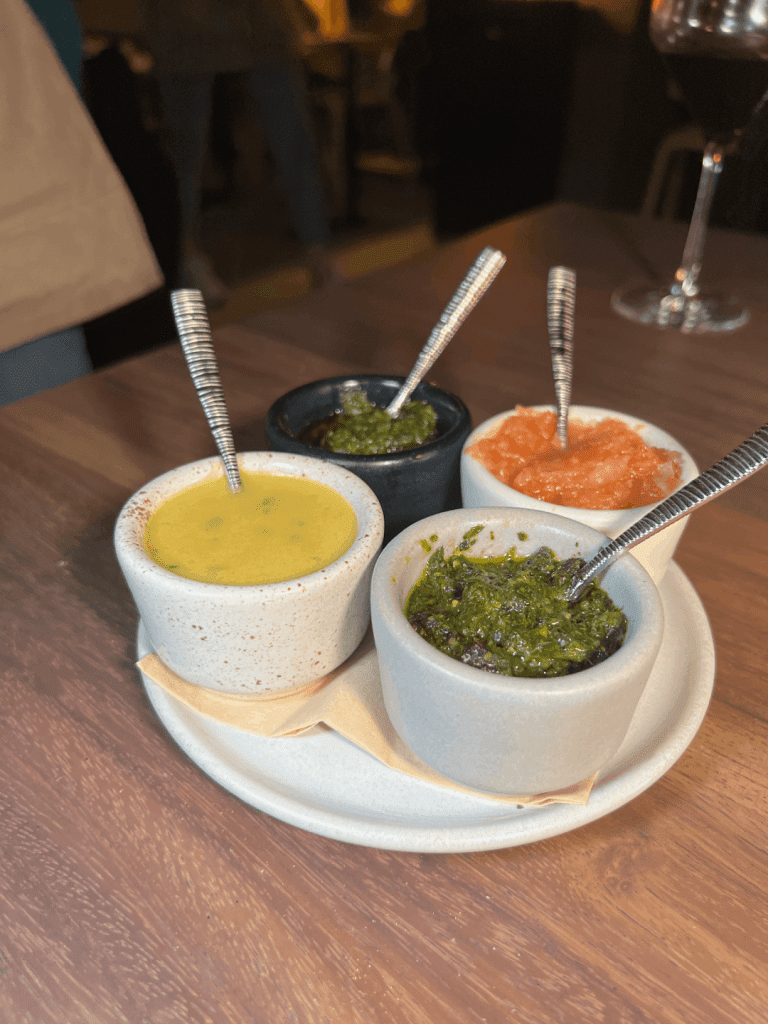 Four small bowls of different sauces with spoons on a wooden table.