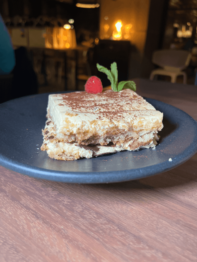 A slice of tiramisu on a dark plate, garnished with a raspberry and mint leaves.