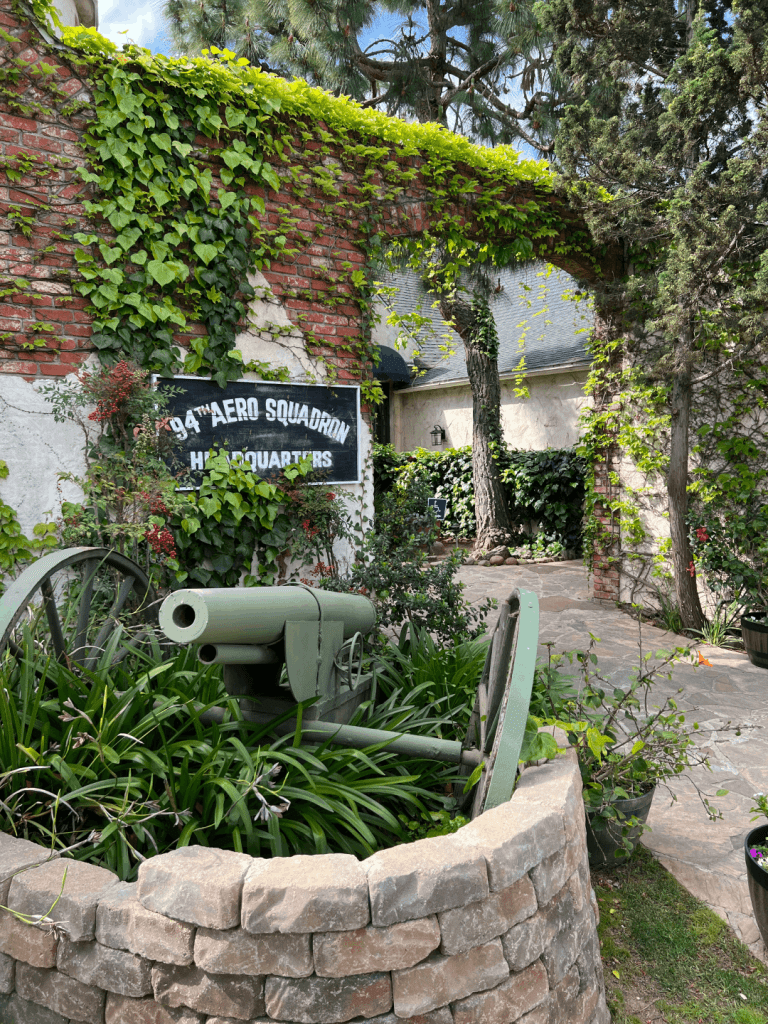 Green cannon surrounded by plants and stone wall, with a brick and ivy-covered wall in the background displaying a "94th Aero Squadron Headquarters" sign.