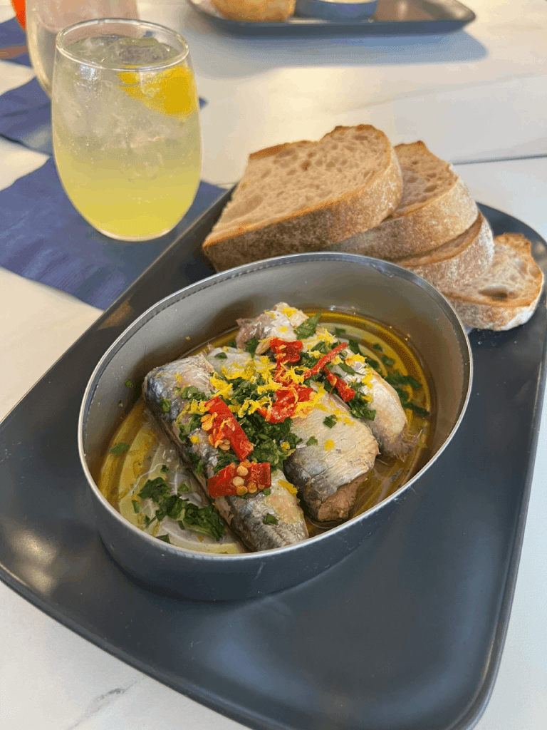 A dish of sardines garnished with herbs, lemon zest, and red peppers, served with sliced bread and a glass of a citrus drink on the side.