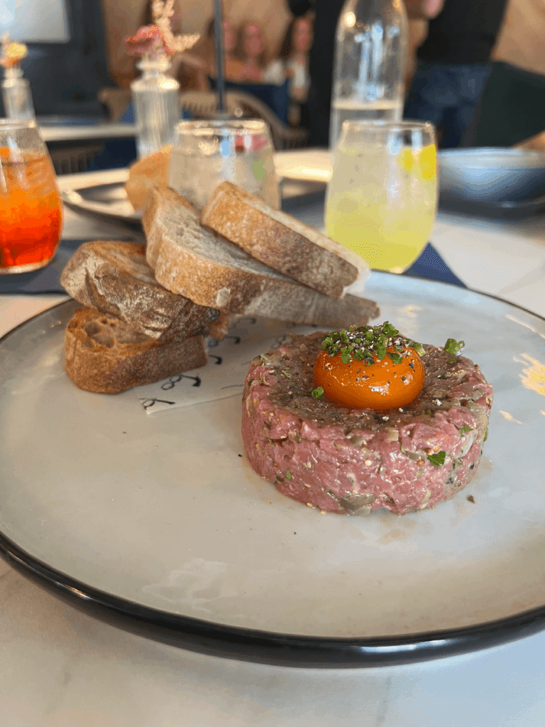 Plate of steak tartare topped with an egg yolk, served with slices of bread, and drinks in the background.