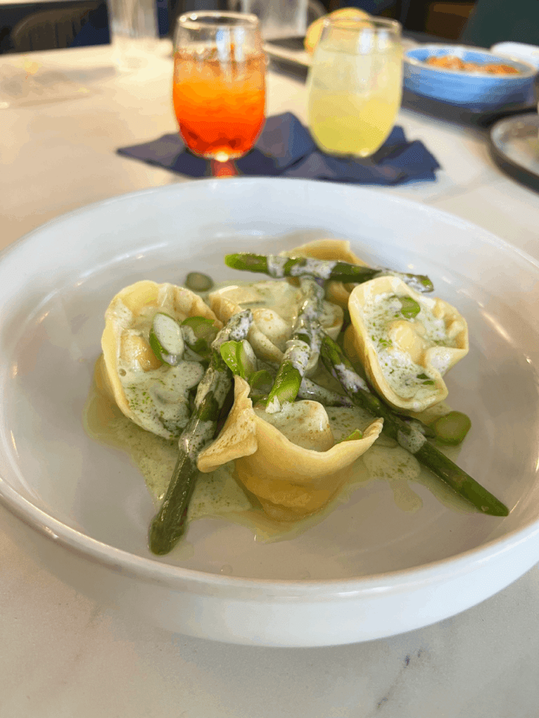 A dish of ravioli with asparagus and creamy sauce, served in a white bowl, with two glasses of colored drinks in the background.