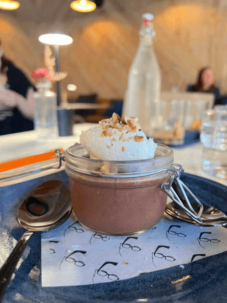 A jar of chocolate dessert topped with whipped cream and nuts, served on a blue plate with a spoon, in a warmly lit restaurant setting.
