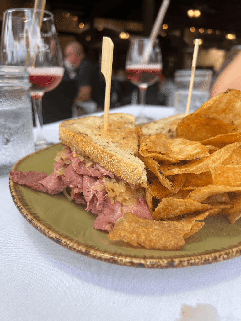 A sandwich with corned beef and sauerkraut on rye bread is served with potato chips on a plate. Two glasses of rosé wine are in the background.