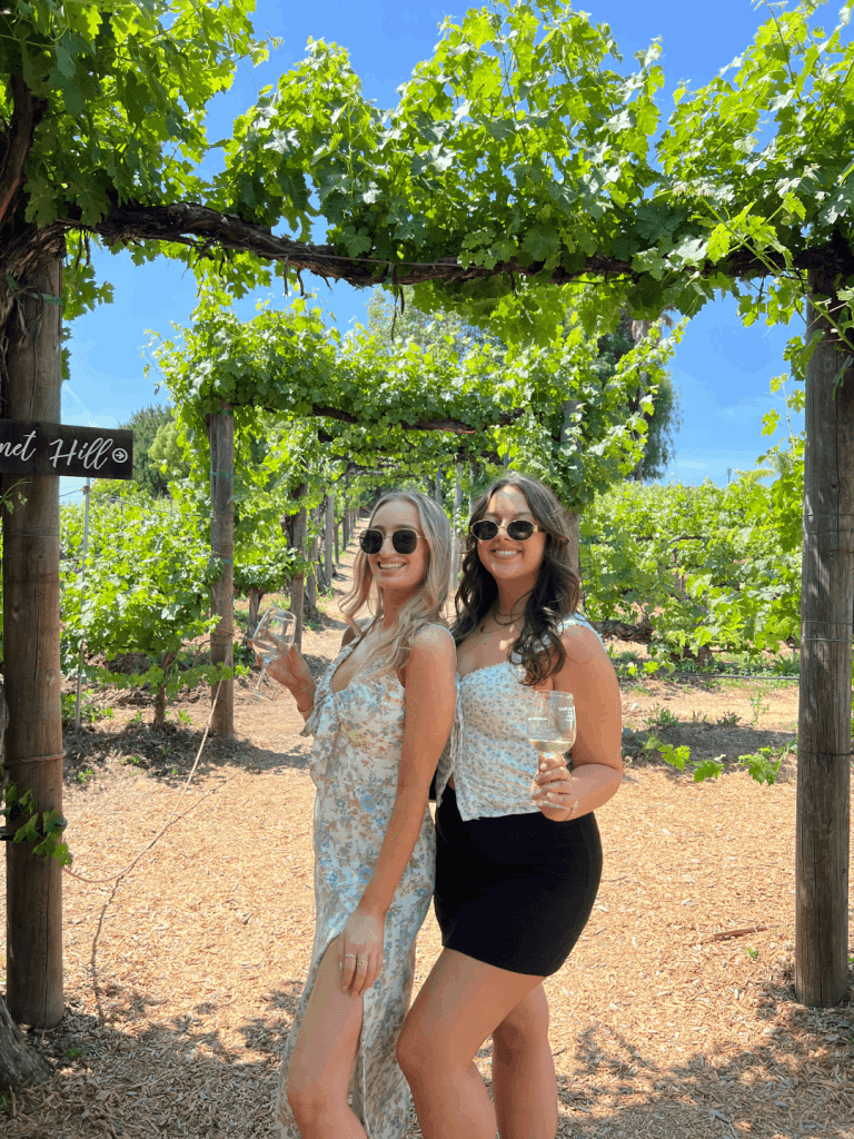 Two women holding wine glasses standing on a leafy pathway in a vineyard.