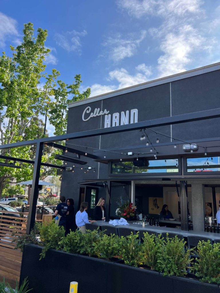 Outdoor seating area of a building with "Cellar HAND" sign, surrounded by greenery, and people standing near a counter under a blue sky.