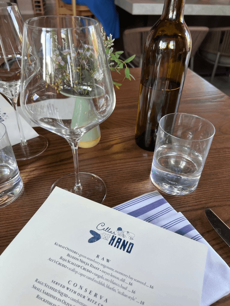 Table setting with wine glasses, a bottle, a water glass, a menu labeled "Cellar Hand," and a small vase with flowers on a wooden table.