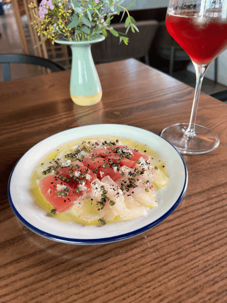 A plate of carpaccio garnished with capers and seasoning, accompanied by a glass of red wine and a vase with flowers on a wooden table.