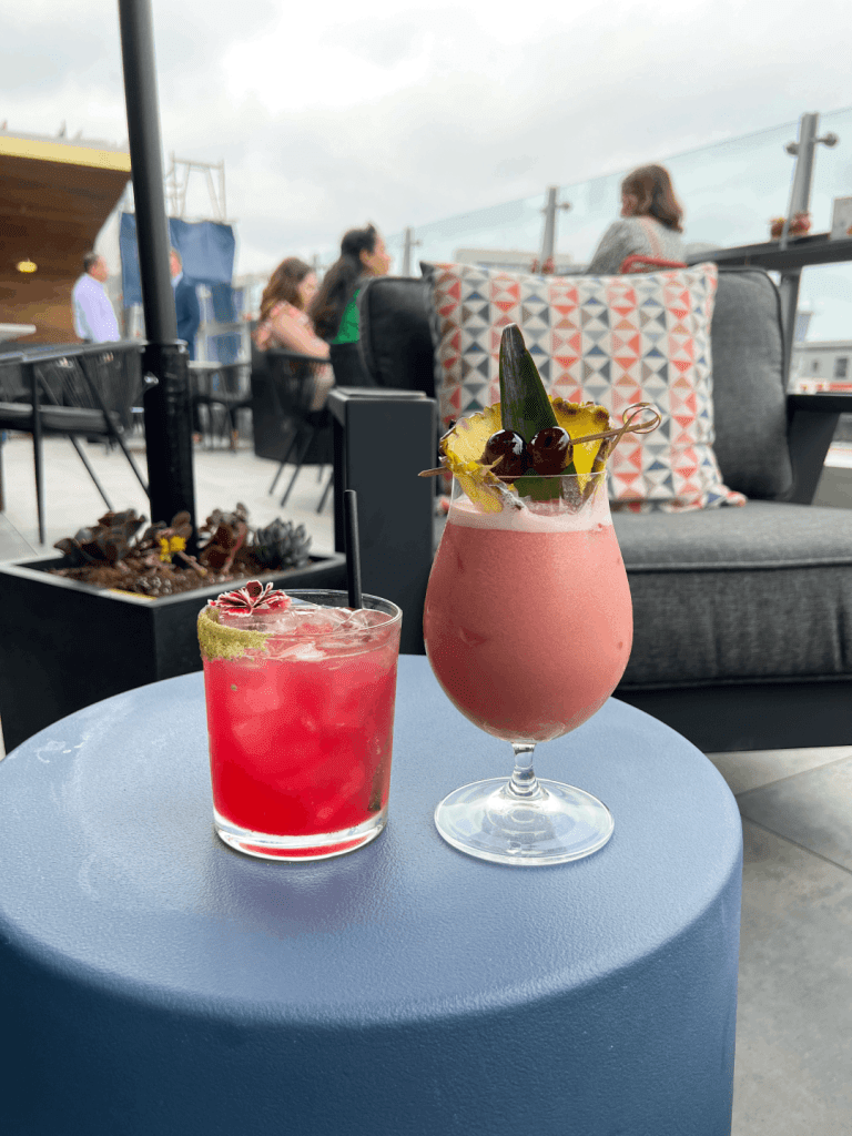 Two colorful cocktails on a blue table, with outdoor seating in the background.