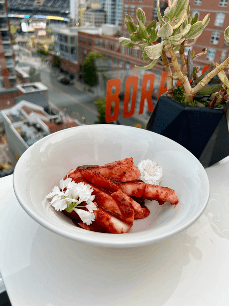 A white bowl filled with marinated sliced octopus garnished with white flowers, set on a rooftop with a cityscape and a small plant in the background.
