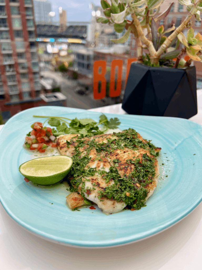 Grilled fish with herbs on a blue plate, garnished with lime, pico de gallo, and cilantro, with an urban background and a potted plant.