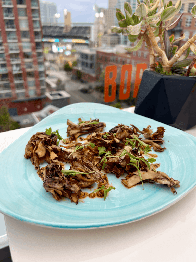 Plate with grilled mushrooms garnished with herbs, placed on a table with a succulent plant and an urban cityscape view in the background.