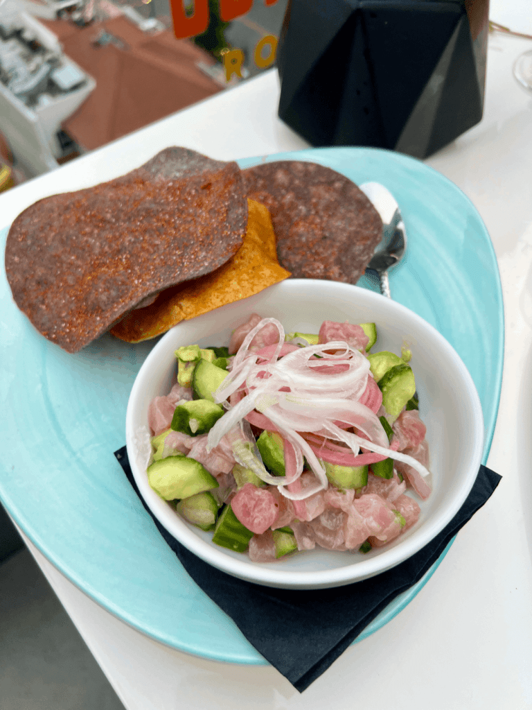 A bowl of ceviche with diced fish, cucumbers, and sliced onions is served with blue and yellow corn tostadas on a light blue plate.