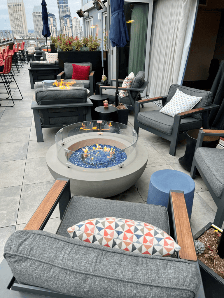 Outdoor patio with gray cushioned seating, colorful pillows, a modern fire pit with blue glass stones, surrounding tall city buildings, and an urban skyline view.