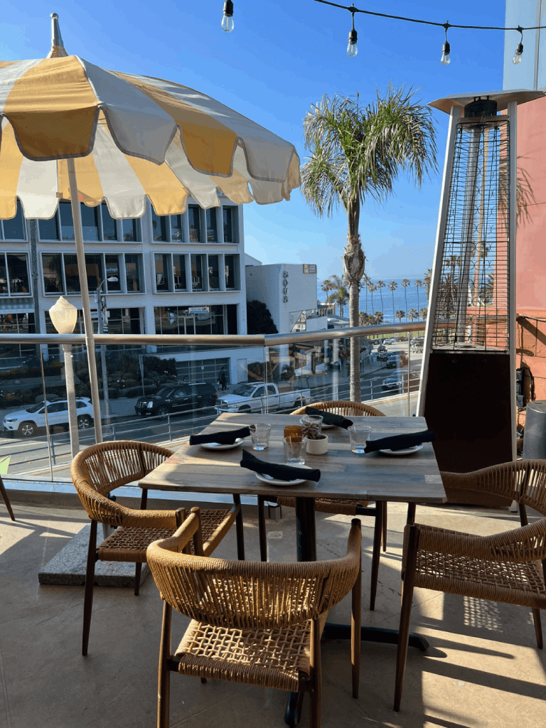 Outdoor dining area with a wooden table and wicker chairs under a striped umbrella, overlooking a street and palm trees with an ocean view in the background.