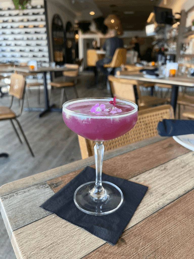 A vibrant purple cocktail garnished with small flowers, served in a stemmed glass on a wooden table with a black napkin in a cozy restaurant setting.