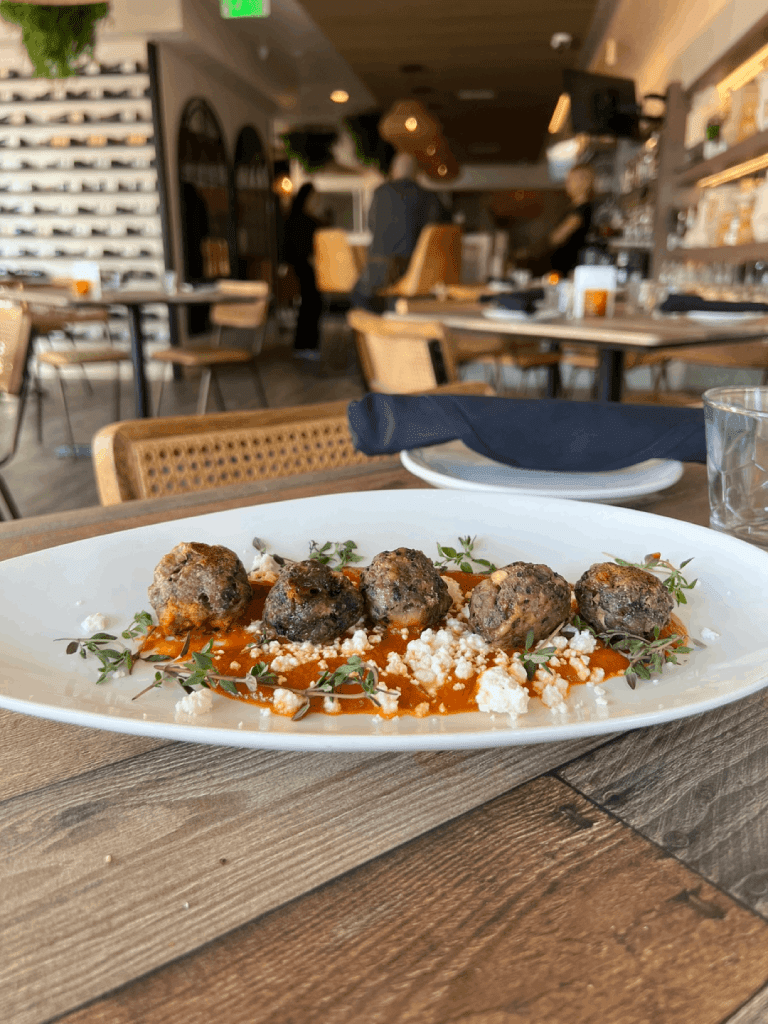 Four meatballs on a bed of tomato sauce with crumbled cheese and herbs, served on a white oval plate in a restaurant setting.