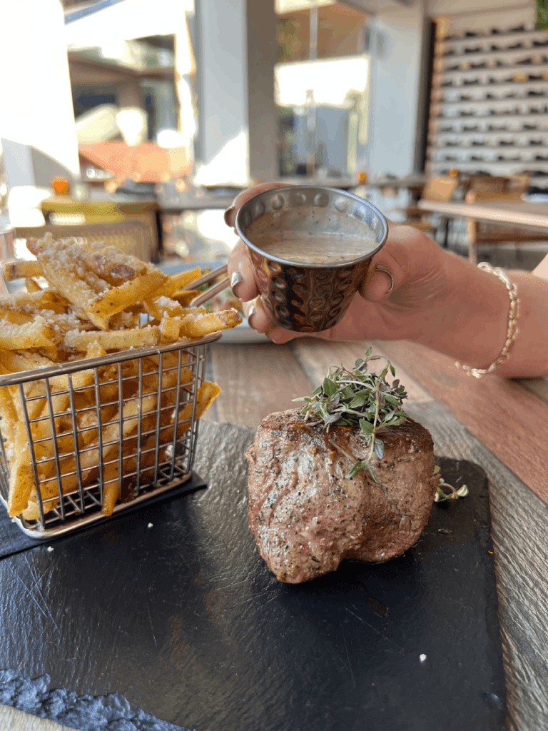 Grilled steak topped with herbs on a slate board, accompanied by a basket of seasoned fries, with a hand holding a metal cup of sauce.