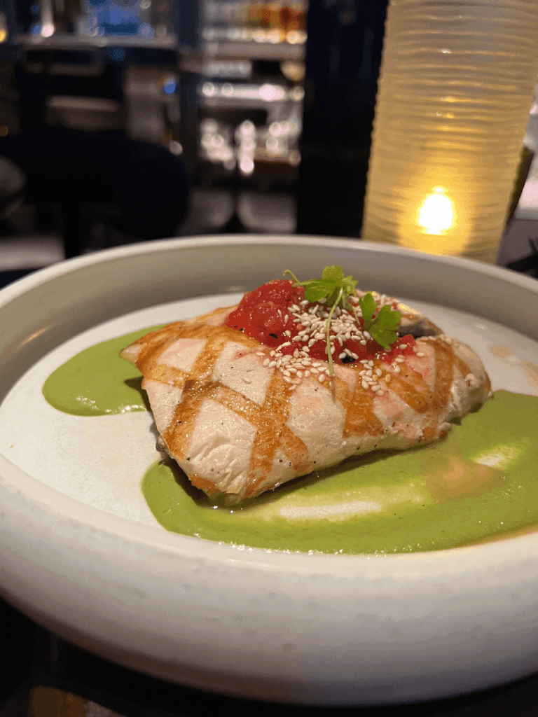 Grilled fish fillet with a green sauce, topped with diced tomatoes and sesame seeds, garnished with fresh herbs in a white dish.