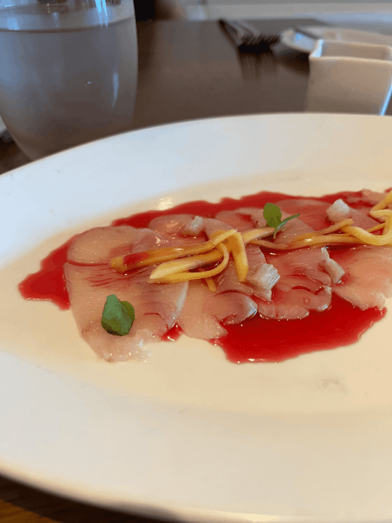 Plate of thinly sliced raw fish with red sauce and garnished with herbs and julienned vegetables.