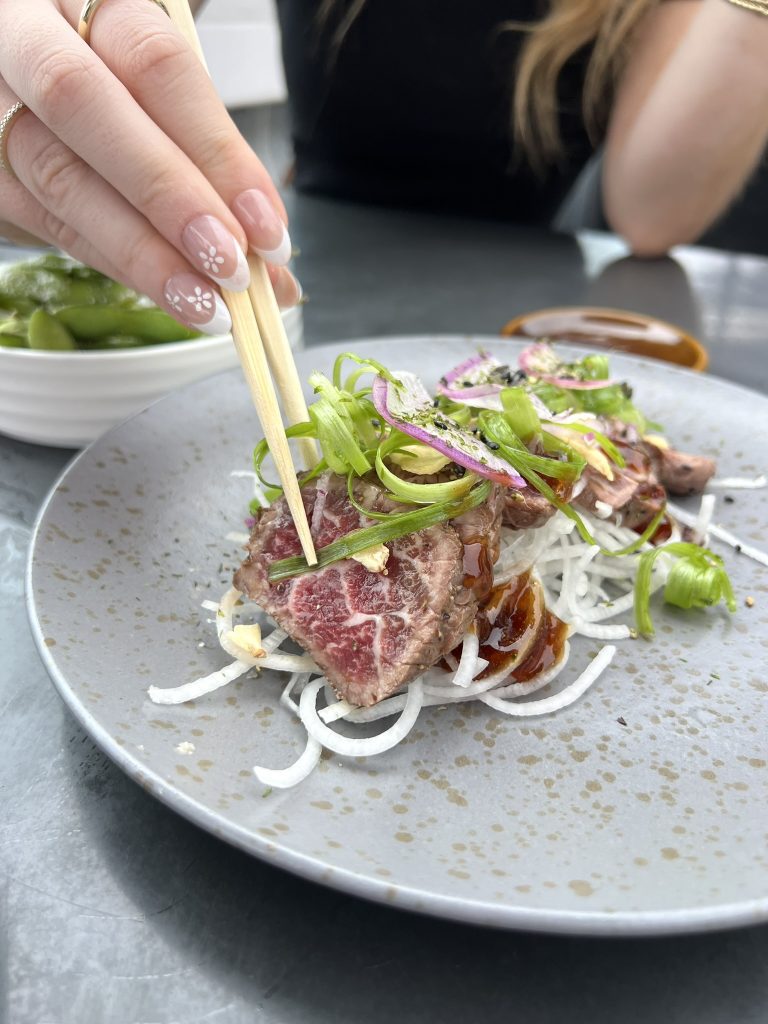 A person using chopsticks to pick up a piece of seared beef on a plate, garnished with sliced onions and green vegetables. Edamame beans are visible in the background.