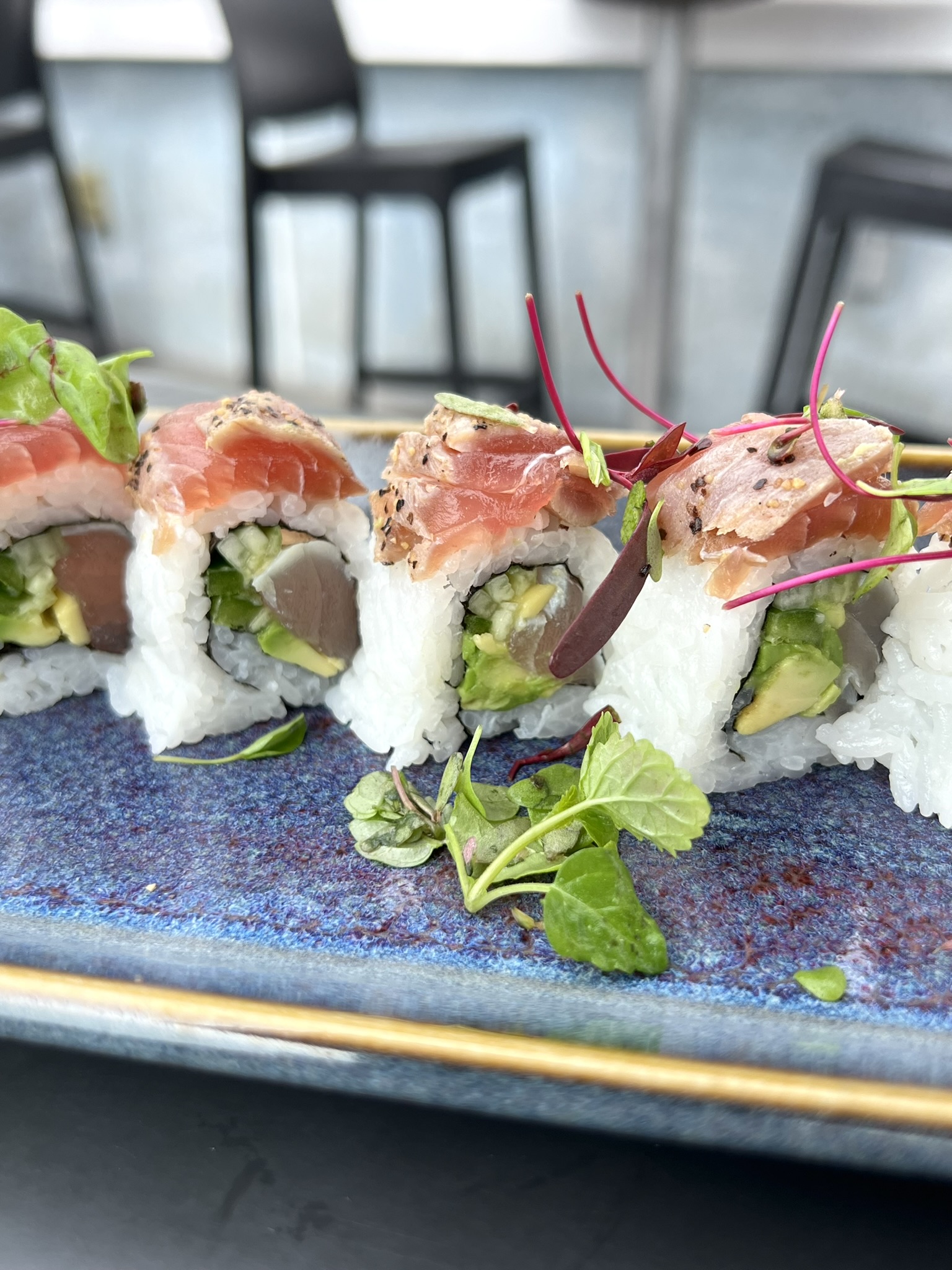 Sushi rolls with raw fish slices on top, avocado, and rice, garnished with microgreens on a blue ceramic plate.