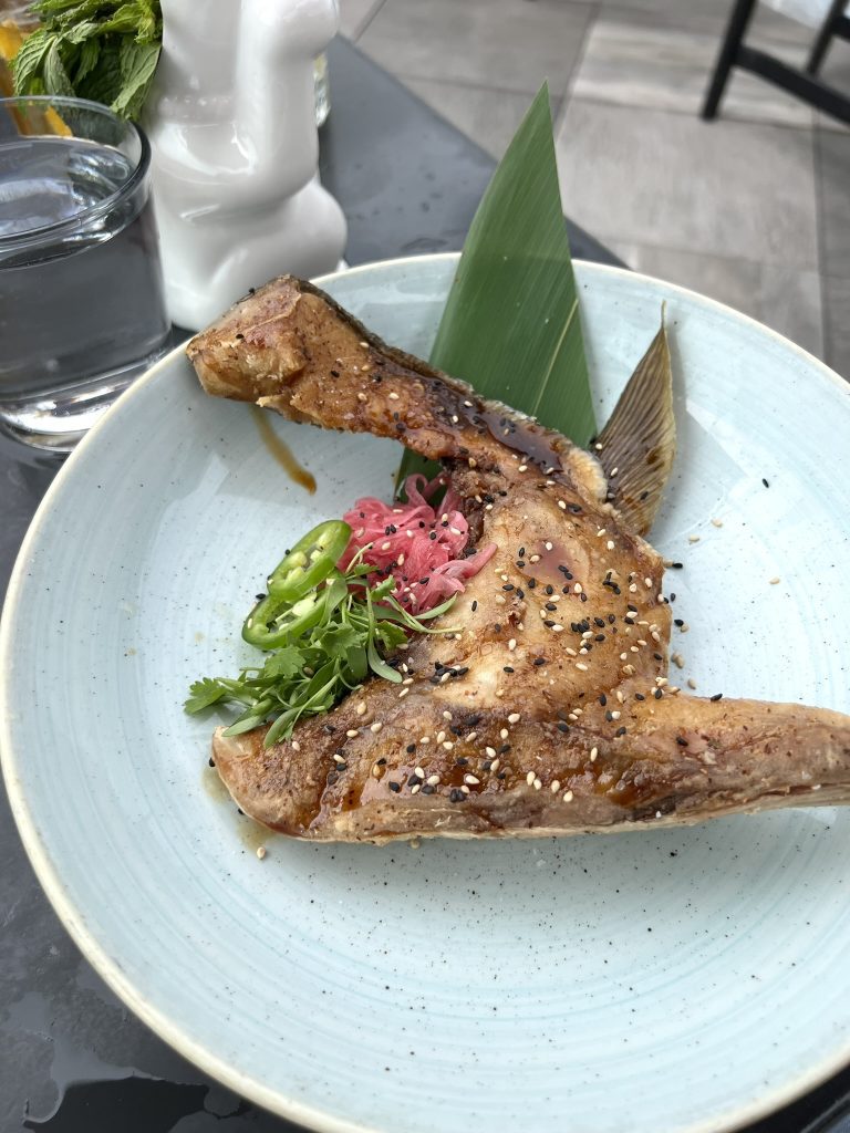 Grilled fish collar garnished with sesame seeds, cilantro, sliced jalapeños, and pickled onions, served on a light blue plate.