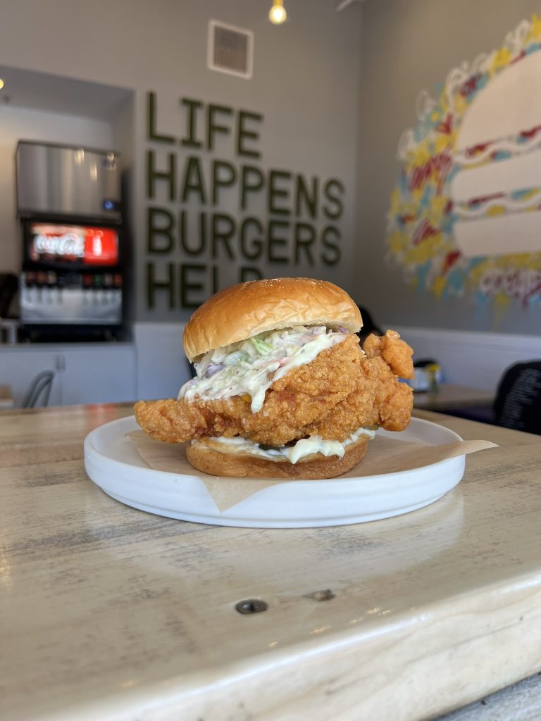 A fried chicken sandwich on a white plate, with "Life Happens Burgers Help" on the wall in the background.