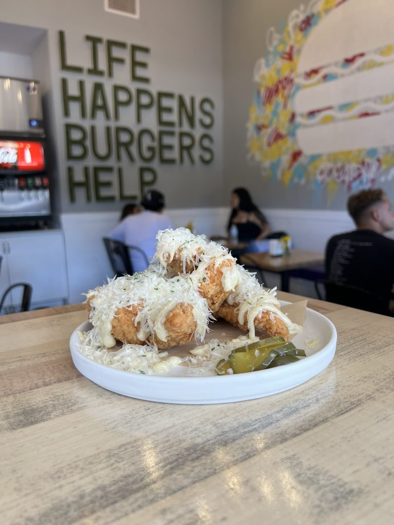 Plate of fried food topped with cheese and a slice of pickle in a diner with "Life Happens Burgers Help" on the wall.